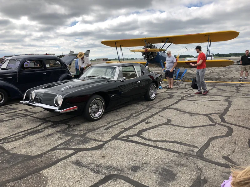 A black Studebaker Avanti with wire wheels, parked in front of a yellow biplane at an airfield show.